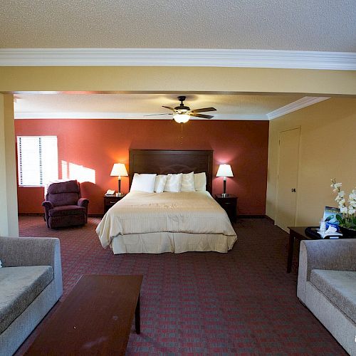 A hotel room features a bed, two couches, a coffee table, a recliner, lamps, and a ceiling fan. The room has warm tones and is well-lit.