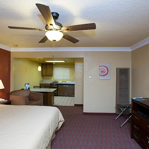 The image shows a hotel room with a bed, nightstand, lamp, TV, dresser, ceiling fan, and kitchenette area with a fridge and microwave in the background.