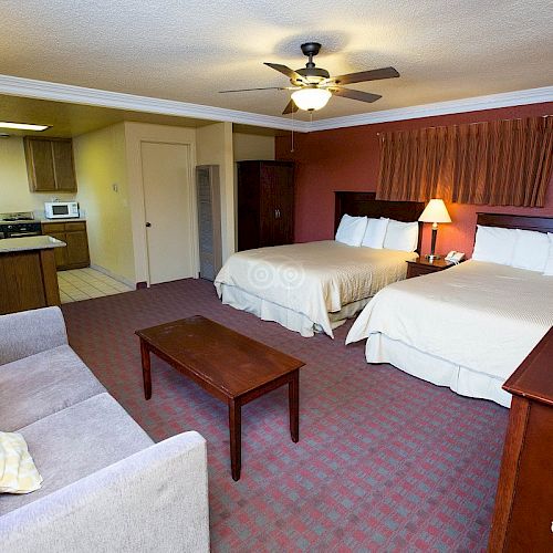 A hotel room with two beds, a sofa, a coffee table, a dresser, and a kitchenette area with a microwave, stove, and a fridge.