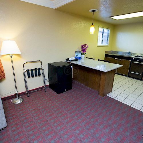 A cozy hotel room with a kitchenette, sofa, lamp, and a small table. The kitchenette has a microwave, stove, and coffee maker.