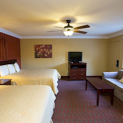 The image shows a hotel room with two beds, a TV, sofa, table, lamp, and a ceiling fan. The decor is simple with beige tones and a wall painting.