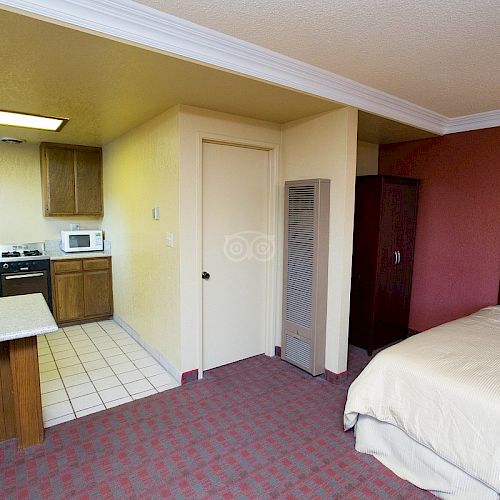 This image shows a hotel room with a bed, a kitchen area with cabinets, a stove, and a fridge, and a small dining counter. The floor is carpeted.