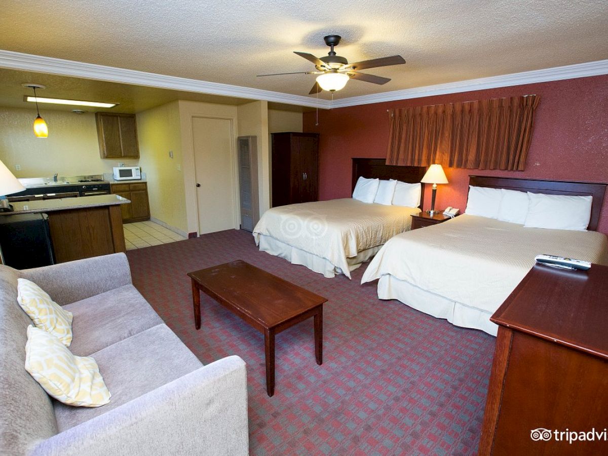 This image shows a hotel room with two beds, a sofa, coffee table, lamp, TV stand, and a small kitchenette area with a microwave, sink, and stove.