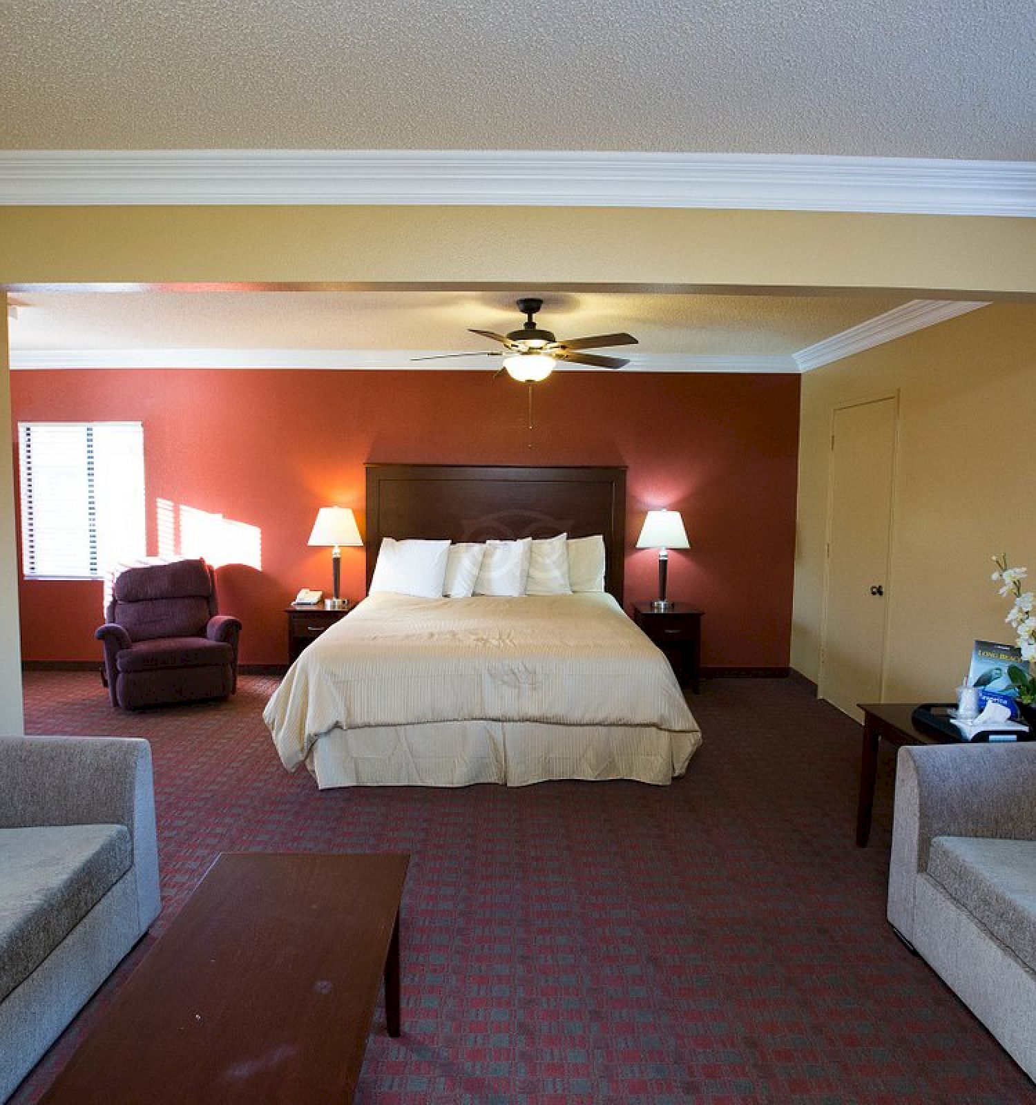 The image shows a spacious hotel room with a bed, two sofas, a coffee table, lamps, and a ceiling fan. It is well-lit with natural light.