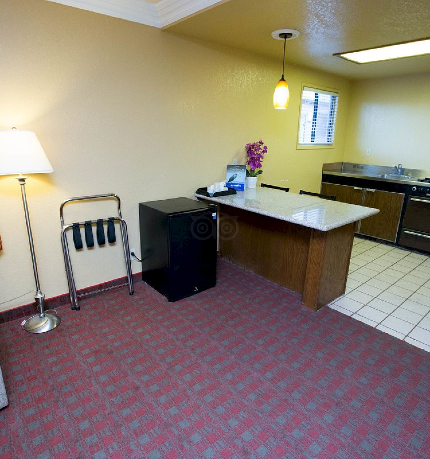 A cozy hotel room with a sofa, lamp, luggage rack, and kitchenette featuring a stove, microwave, and refrigerator.