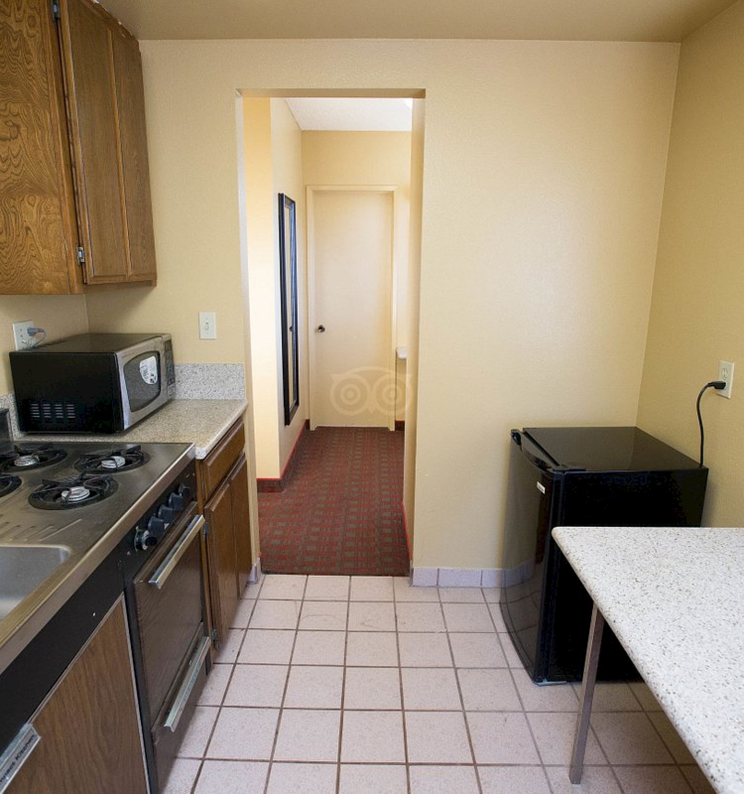 A small kitchen with a sink, stove, microwave, dishwasher, refrigerator, table, and cabinets. The kitchen opens into a hallway with a carpet.